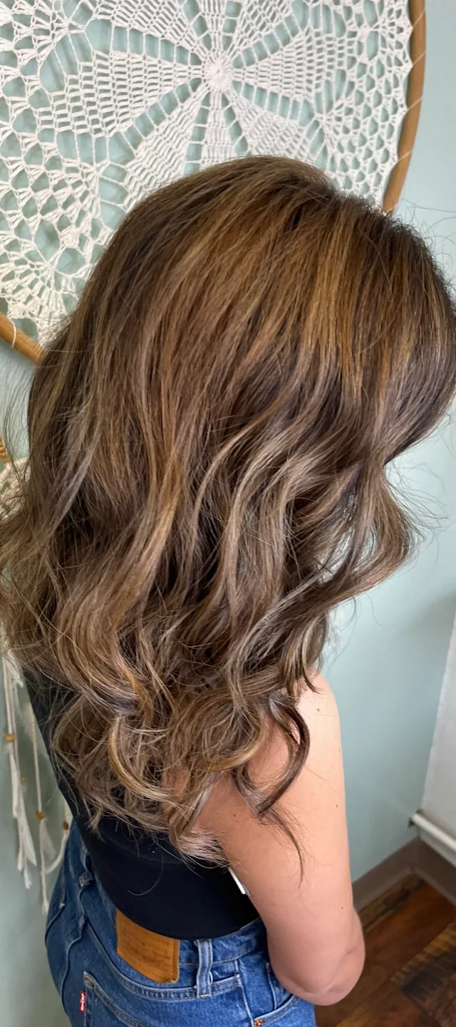 A woman seen from the side-back, showing her long, wavy brown hair with warm-toned highlights. A white crocheted dreamcatcher hangs on the light-colored wall behind her.