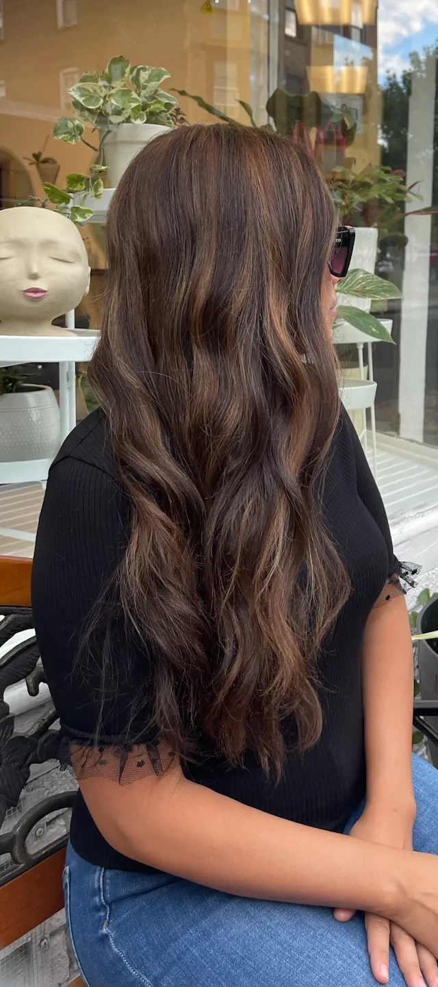 A woman with long, wavy, dark brown hair with subtle highlights sits on a bench, seen from the side-back. She is wearing a black shirt and jeans, and through the window behind her, plants are visible.